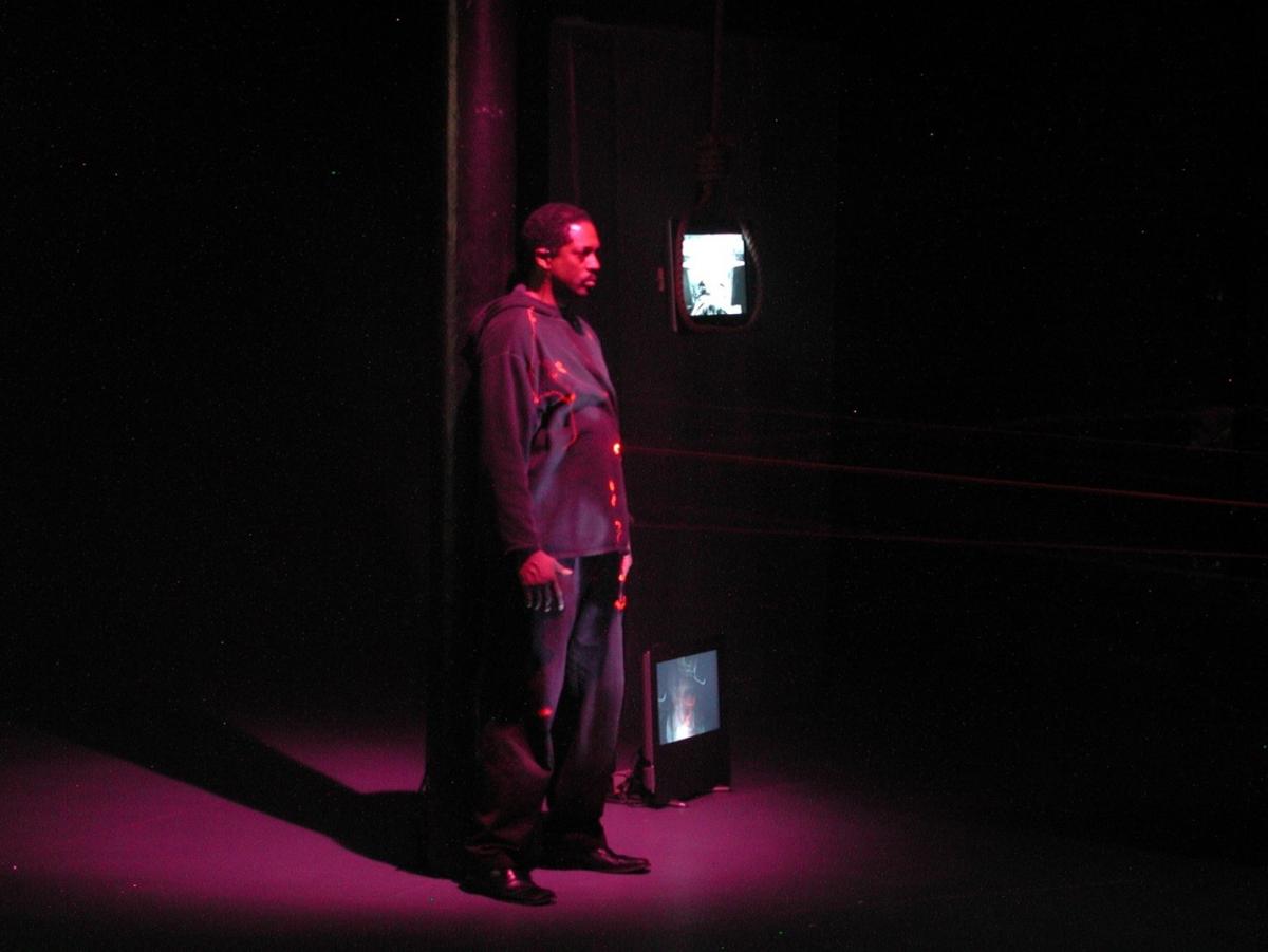 A person in red light stands on a stage, hands placed firmly on their thighs. There appears to be a small screen displaying an image next to their legs.